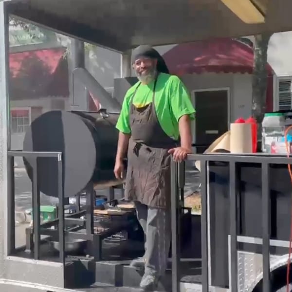 Man in a green shirt and apron standing next to a large black smoker on a food truck.