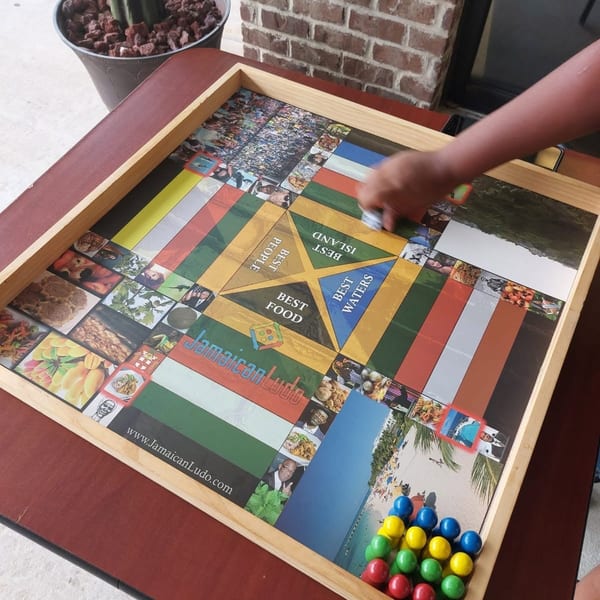 A person's hand plays Jamaican Ludo on a wooden board with colorful game pieces.