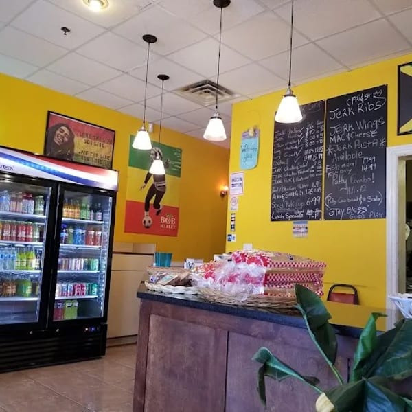 Interior of a vibrant Jamaican restaurant with yellow walls, a menu board, and a drink cooler.