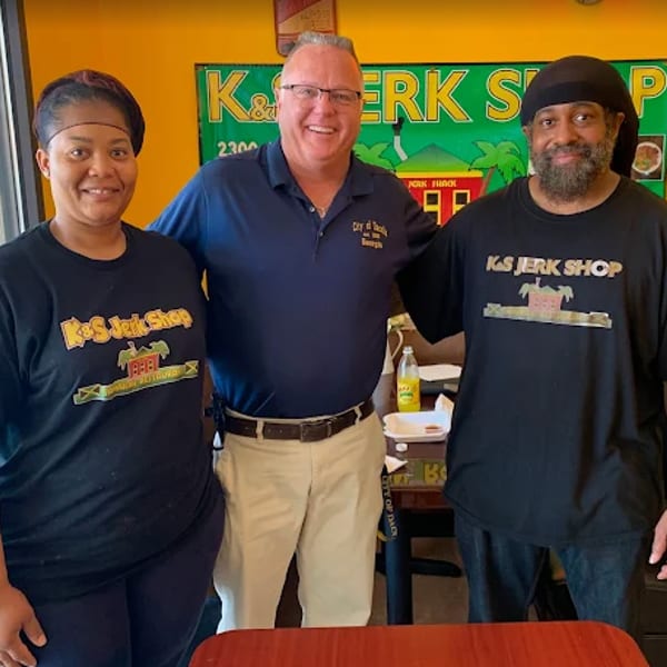 Three people, two wearing K&S Jerk Shop shirts, stand smiling in front of a colorful sign.