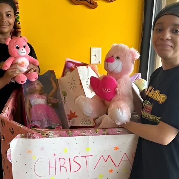 Two young women with a box of toys, including pink teddy bears and a doll, for Christmas.