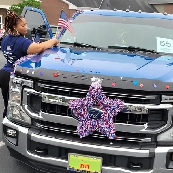 A woman decorates a blue Ford truck with patriotic garlands and an American flag.