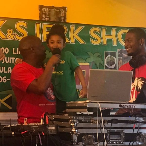 A man holds a child singing into a microphone, with a DJ smiling behind a laptop.