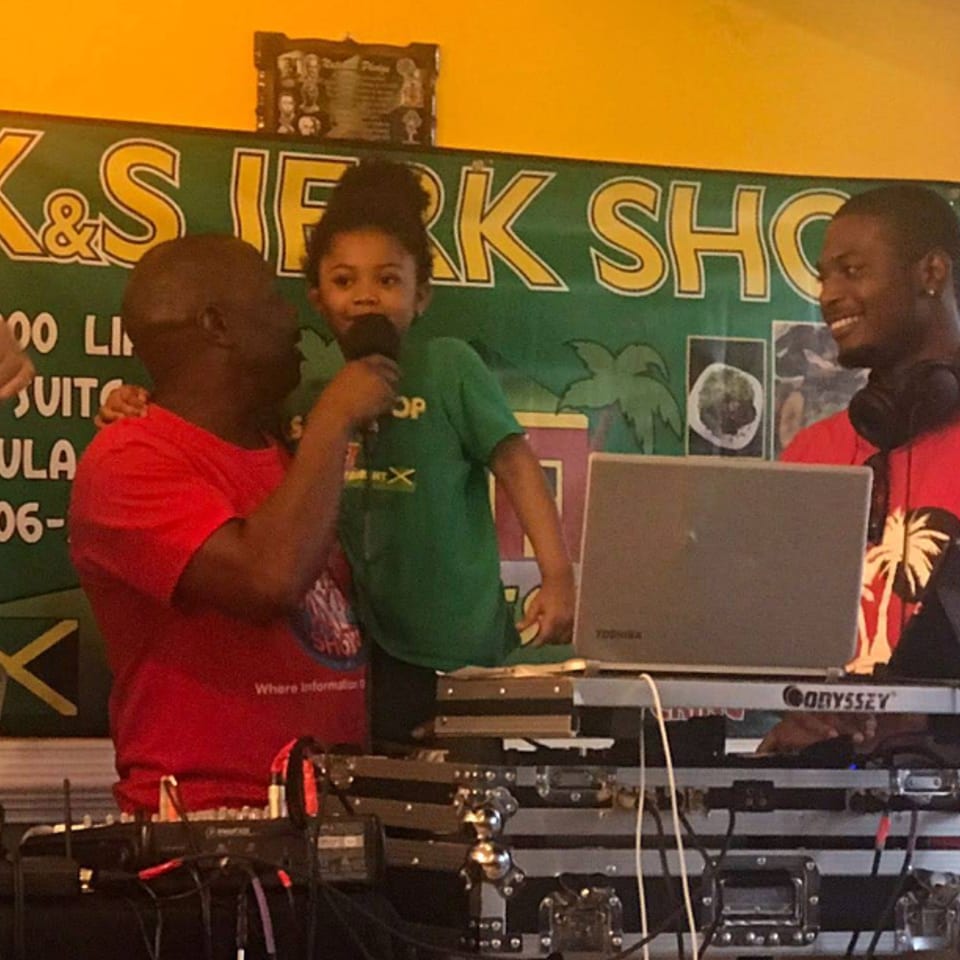 A man holds a child singing into a microphone, with a DJ smiling behind a laptop.