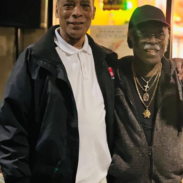 Two men smiling, one in a white polo and black jacket, the other in a black cap and gold chains.
