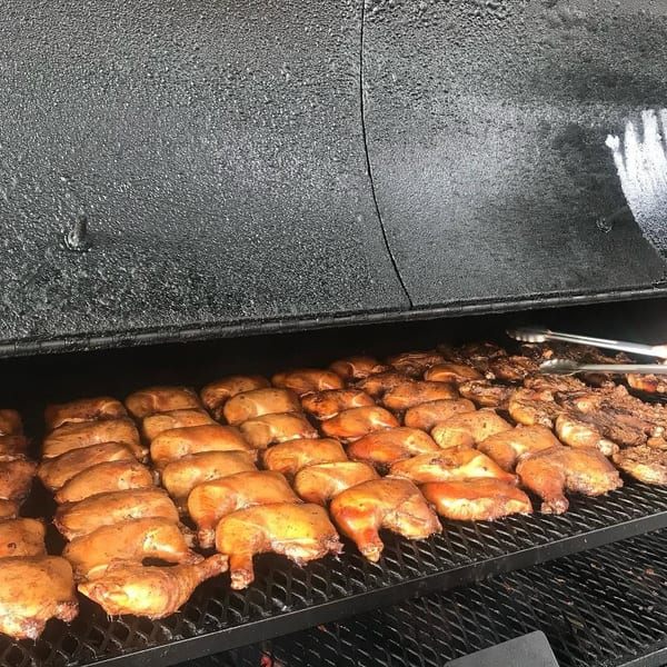 Rows of chicken quarters and other meat cooking on a large black grill, with tongs turning food.