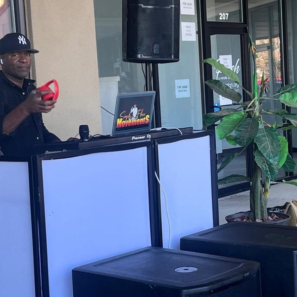 A DJ in a black shirt and Yankees cap holds red headphones, standing behind a Pioneer DJ setup.