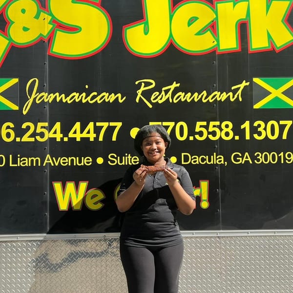Smiling woman holding a piece of jerk chicken in front of the &S Jerk Jamaican Restaurant food truck.