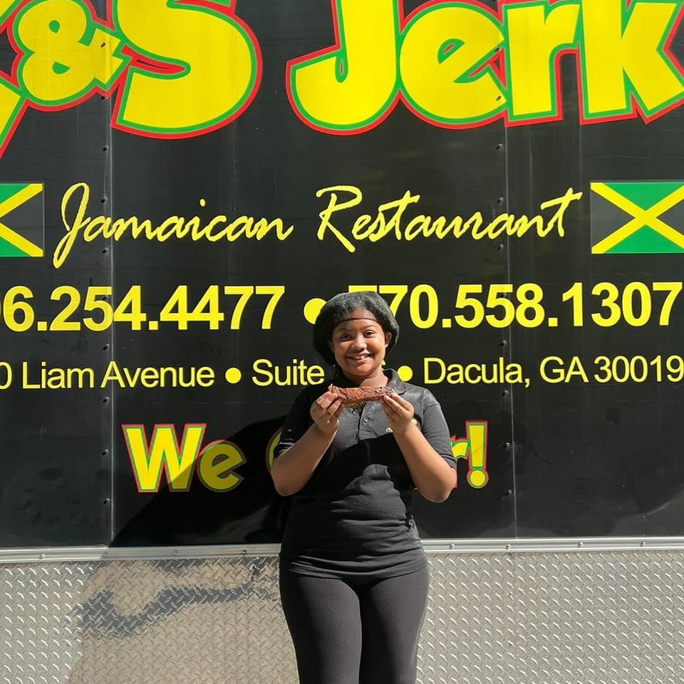 Smiling woman holding a piece of jerk chicken in front of the &S Jerk Jamaican Restaurant food truck.