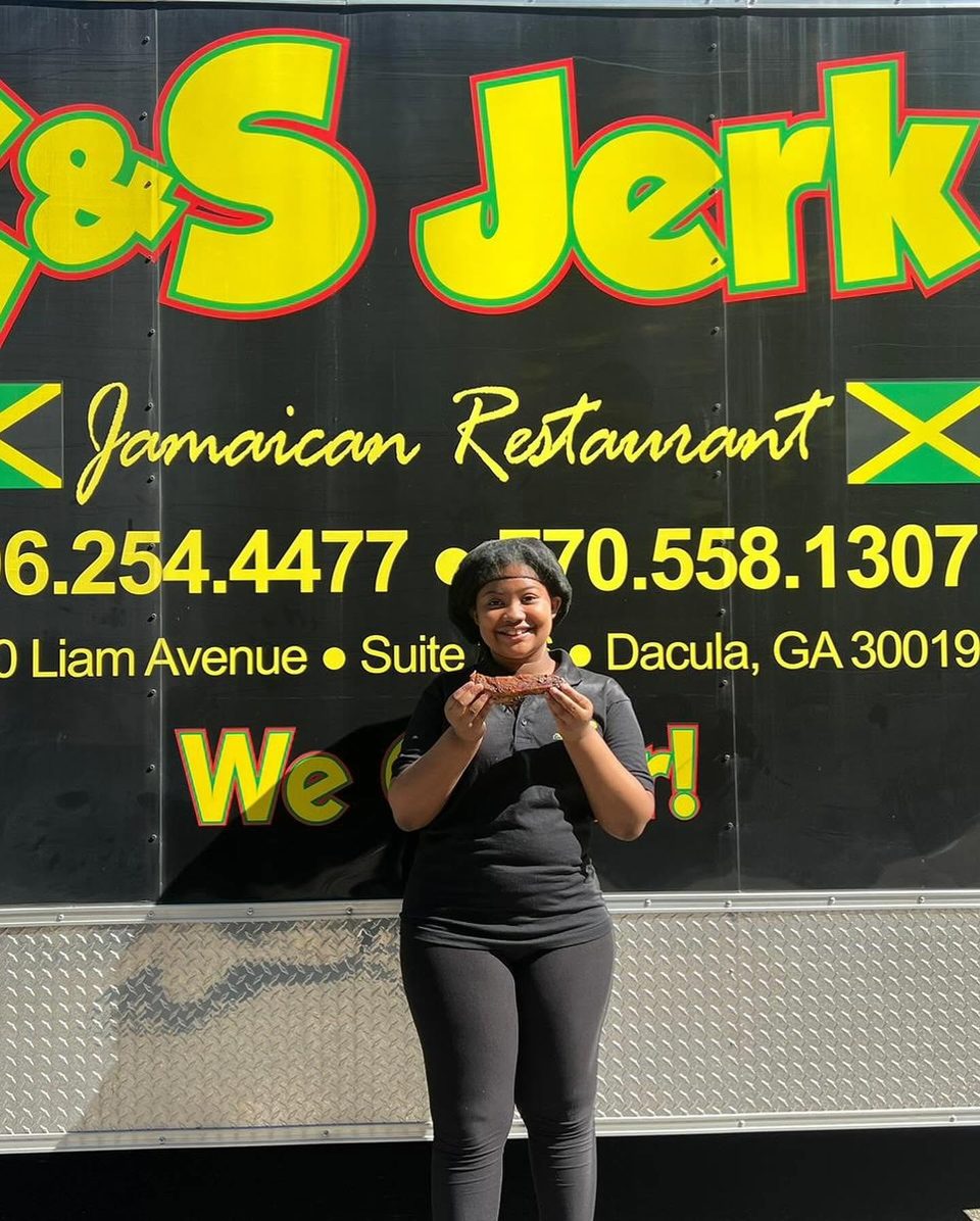 Smiling woman holding a piece of jerk chicken in front of the &S Jerk Jamaican Restaurant food truck.