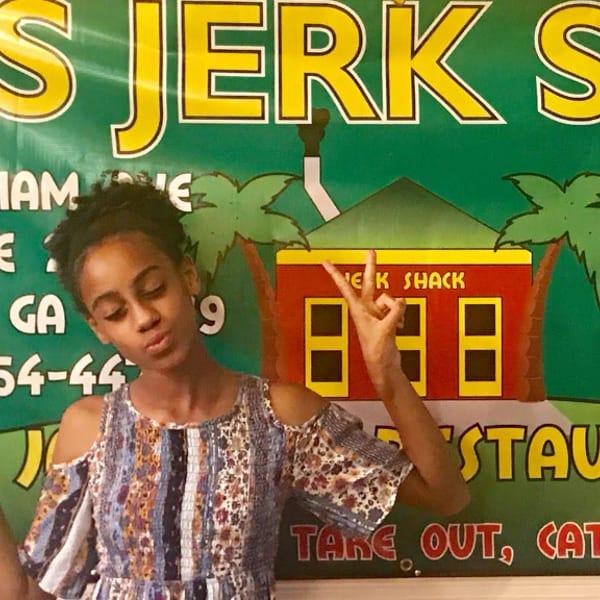 A young girl poses with peace signs in front of a green "Jerk Shack" restaurant banner.