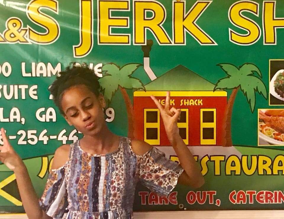 A young girl poses with peace signs in front of a green "Jerk Shack" restaurant banner.
