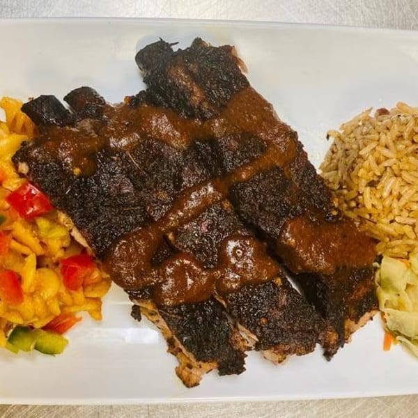 Grilled ribs with sauce, pasta salad, rice, and cabbage on a white rectangular plate.