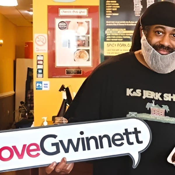 Man in a beard net and "K&S Jerk Shack" shirt holds a food container and #LoveGwinnett sign.