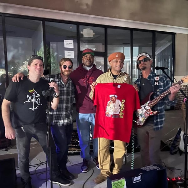 Five men, some holding instruments and a red t-shirt, pose together outdoors at night.