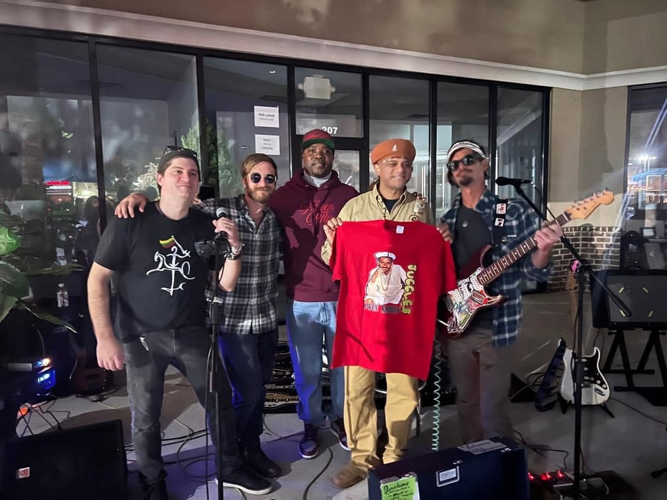 Five men, some holding instruments and a red t-shirt, pose together outdoors at night.