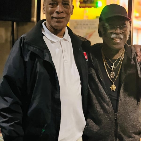 Two men smiling, one in a black jacket and white polo, the other in a black cap and gold chains.