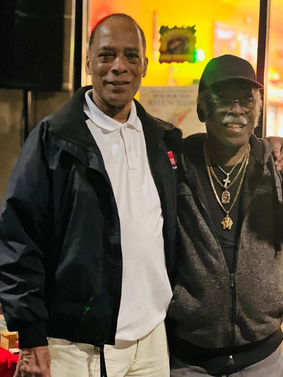 Two men smiling, one in a black jacket and white polo, the other in a black cap and gold chains.