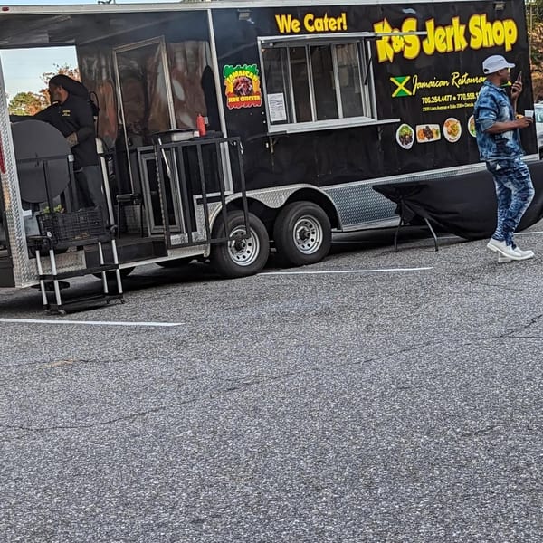 A black K&S Jerk Shop food truck with a person cooking and another walking by.