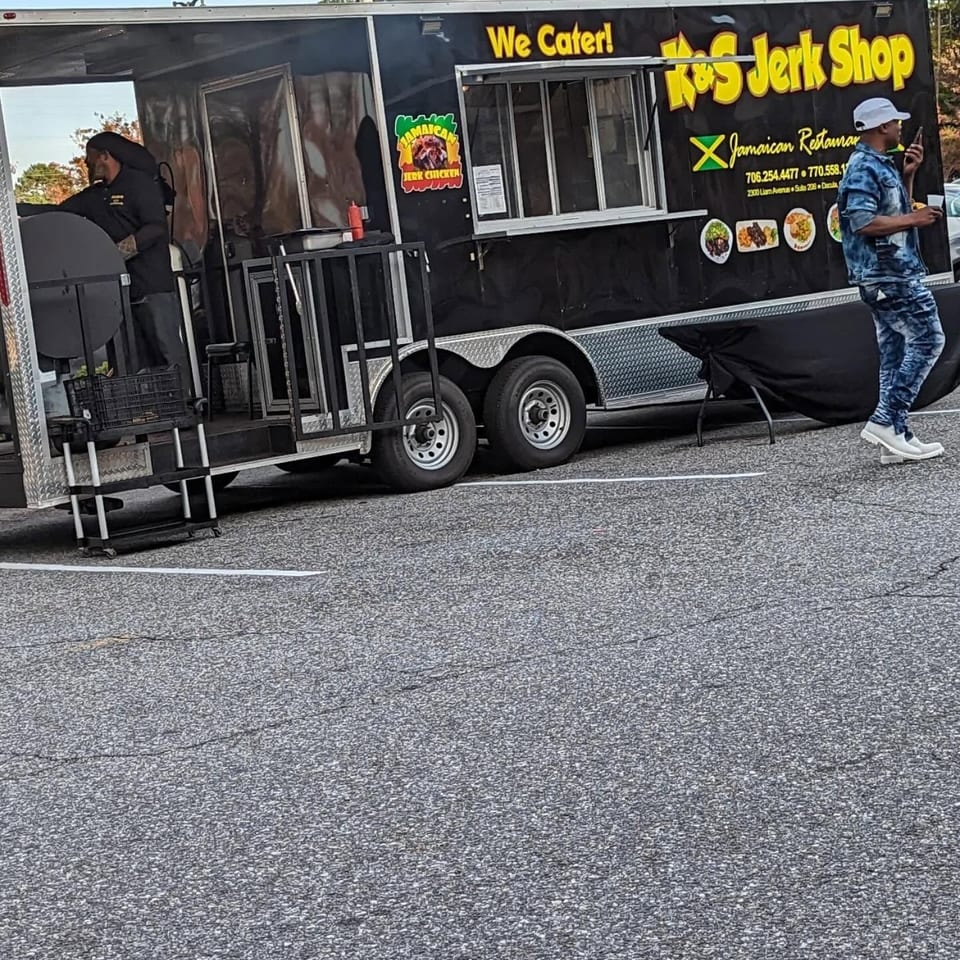 A black K&S Jerk Shop food truck with a person cooking and another walking by.